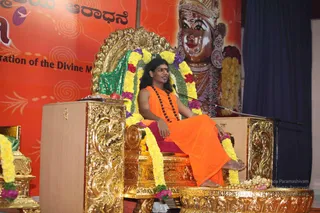 A seated man in saffron-orange robes and a dark rudraksha mala sits on an ornate gold throne decorated with yellow and purple flower garlands and green fabric, speaking at a microphone on a stand. Behind him a large red banner shows a golden deity murti with a crown and earrings, bordered by a Kannada-script heading and an English line about a celebration of the Divine. A matching gold-and-flower seat stands at the left edge. The setting is a stage with a dark blue curtain. Swamiji (probable): the seated figure has long swept-back hair, saffron clothing, a rudraksha mala and a gold throne with garlands, presented as the speaker giving a discourse.