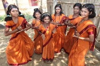 A photograph of six young girls standing outdoors in a group, posed closely together with their hands gesturing in front of them. All wear orange saris with maroon borders and rudraksha beaded necklaces, with white and red forehead markings. Some wear hair bands or hair ornaments. Behind them are a bamboo fence, a red wall and green foliage, with dappled sunlight on the ground. No a person candidate is visible.