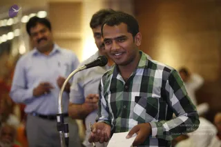 A smiling young man in a green, white and black checked button-up shirt stands at a gooseneck microphone, holding a folded paper and a pen in his hands. Behind him, two men in light collared shirts stand in a softly lit hall, one with a moustache and the other holding a paper. Blurred seated audience members fill the lower foreground and right edge.