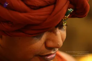 Close-up photograph in profile of a man wearing a rust-orange turban wrapped in folds, with a green and gold jeweled ornament tucked at the temple. His eyes are lowered and he appears absorbed. The warm, soft-focus background is golden. Appearance cues (turban, orange clothing, ornate adornment) are consistent with a person candidate but the face and details are not clear enough to confirm, so identity remains uncertain.