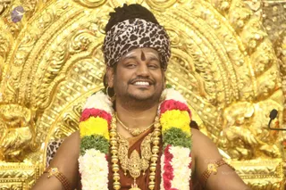 A smiling man sits in front of an ornate gold repoussé backdrop, seated on a throne-like seat. He wears a leopard-print turban-style head wrap, a tilak on his forehead, and a red-brown garment. He is adorned with a multi-strand gold necklace featuring a deity pendant, gold armlets, and a garland of red roses, yellow marigolds, green leaves and white flowers draped over his shoulders. A microphone stands at the right edge of the frame.