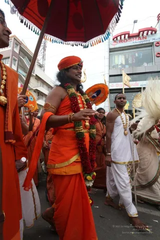 Street procession in a city with a red-and-orange shopping mall behind it. A man in the centre wears an orange turban with a small ornament, orange robes, a thick red and green flower garland and gold ornaments on his upper arm and wrists. He walks under a large red umbrella with tasselled fringe held by a man on the left in orange robes and gold necklace. Behind him, a man in plain white dhoti and gold chain necklace carries a white fly-whisk-style staff. Several people in orange and cream garments follow, and an orange ceremonial fan and gold standard are visible. The procession is a street festival walk, with a hand held out in blessing. Visible people: about four or five in the foreground and several more behind.