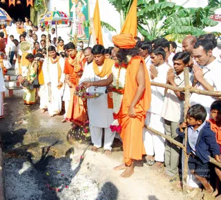 A photograph of an outdoor temple procession with a fire-walking area in the foreground, where ash, embers and scattered flower petals lie on the ground. A central figure in a saffron-orange robe and matching orange turban, with long dark hair and a white-and-pink flower garland, holds a brass tray of flowers and gestures over the fire path. Around him, men in white dhotis and white shirts, some with orange shawls, stand and watch; a few people on the right have their palms pressed together in namaskar behind a wooden railing, and a young boy in a blue jacket leans on the rail. Behind them a painted deity banner, orange flags and striped ceremonial umbrellas are visible, and a drummer in yellow stands at left. Appearance is consistent with the page's claim of a person, but the face and identity cannot be confirmed from appearance alone.