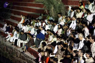 Night outdoor gathering of a large seated crowd on red-brick terraced steps beside a palm tree, with a dark pool or water area at left. Many people sit closely in rows, mostly in white and cream clothing, with some in orange, saffron, blue and green garments; several wear lanyards and some women wear sarees and salwar kameez. Some attendees sit with palms joined or eyes closed in meditation or satsang, while others look toward the stage. A few faces are lit by soft evening light. The scene is a devotee gathering, not a single identifiable leader in clear view.