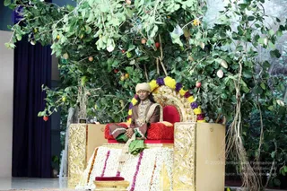 A photograph of a single seated man on an ornate gold-and-red throne (peetha) decorated with marigold, jasmine and multicoloured flower garlands, set beneath a leafy tree hung with fruit. He wears a cream turban, a brown embroidered robe with a gold-toned necklace, and holds a green leaf-wrapped object in his lap with eyes closed. A dark blue curtain hangs at left and a floral carpet of white and purple blossoms lies before the throne. The scene suggests a satsang or darshan setting.