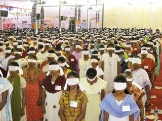 A large indoor hall filled with hundreds of women and girls standing in dense rows, nearly all wearing white cloth blindfolds over their eyes. Many wear saris and salwar kameez in red, orange, yellow, pink, green and blue, with some in white. A few wear small name badges. Red carpet runs along the floor at the right, with plastic water bottles near the front. Overhead hang strings of triangular pennants and tall decorated pillars, and banners are visible on the back wall. No a person figure is clearly visible in the scene.