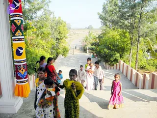 A photograph of villagers walking down a wide concrete path flanked by white-and-red striped low walls, lined with green trees and shrubs under bright daylight. In the foreground a woman in a white polka-dot sari carries a small child on her hip, and beside her a young girl in a green salwar with a yellow dupatta presses her palms together. Behind them a woman in a red blouse and sari, another in a pale pink sari carrying a baby, and several children and men in light shirts walk along the path. At the left edge a tall white pillar is wrapped in a painted textile with bird and floral motifs in red, yellow, blue and orange. A small girl in a bright pink dress stands at right. A distant gate and a motorcycle appear on the road ahead.