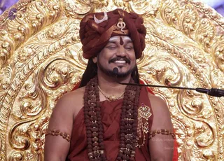 A photograph of a smiling man with closed eyes seated before an ornate gold repoussé throne backdrop. He wears a maroon-red turban with a crescent and a small ornament at the front, red-brown robes, dense rudraksha malas across his chest and shoulders, a gold necklace, a gold brooch with pearls and red stones, and gold-and-bead armlets. A red tilak marks his forehead, and a microphone stand is in front of him. a person, seated in a ceremonial discourse setting with a microphone, shown in saffron-red and rudraksha attire against a gold throne.