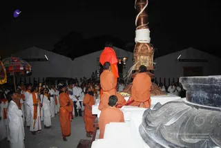 Night photograph of a temple courtyard with a tall copper-and-gold ritual pillar wrapped in white cord at right. A figure in a long orange robe with a red-and-saffron shawl and beaded necklace stands at the pillar's upper base, raising a lamp, while several saffron-robed attendants in orange shawls kneel or stand on a white stone platform. Devotees in white dhotis and shirts stand in rows at left, some with folded hands, one holding a long golden staff; a colourful fringed parasol is at far left. White-walled buildings with signboards frame the background under a dark sky.