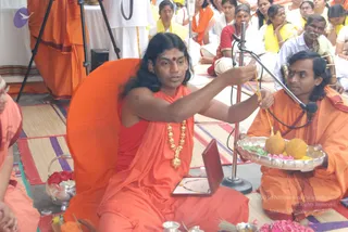 A photograph of a long-haired man in saffron-orange robes seated on an orange cushion, holding a yellow-threaded cord up toward a microphone stand while a man beside him in orange holds a silver tray with fruit and flower offerings. The seated man wears a gold necklace with pendant and a red bindi. Behind them, rows of seated devotees in yellow and orange sashes and white clothing fill the floor, with a woman in a red sari among them. A small wooden box, flowers and brass vessels sit on woven mats in the foreground. a person candidate with long swept hair, saffron robes and gold ornament is present in person, and the page labels the event as a Bidadi temple ritual session.
