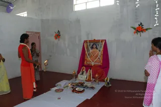 Photograph of a bare-walled hall with a red-brown floor where devotees perform puja before a framed portrait of a long-haired figure in orange robes with a rudraksha mala, garlanded with white flowers and set on a magenta cloth. A woman in an orange top with a white flower garland stands at left holding a lit oil lamp toward the altar. Two other women stand nearby, one in a yellow sari at left edge, one in a pink polka-dot sari with hands folded in prayer at right. The altar on a white cloth holds brass lamps, a kalasha-style vessel, flowers, fruit and a thali. Orange flower decorations and paper garlands hang on the walls.