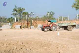 A dusty construction site at an ashram under a clear pale sky. A red tractor with a green canopy pulls a green trailer marked with the letters M and M across bare, reddish-brown earth. Behind a wire fence stand concrete blocks, a pile of gravel, cut stone and red earth banks, with palm and other trees beyond. One person sits on the tractor seat driving. No a person candidate is visible; the scene shows only the tractor operator in a work setting.