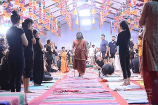 A photograph of a large hall with a striped multicoloured mat runner down the middle and colourful paper lanterns and banners hanging from the ceiling. A central man with long dark hair and a beard walks barefoot in a rust-orange robe with a gold-edged cloth wrap and carries a walking staff; the page indicates he is a person, though his appearance is not conclusive enough to confirm it. Around him, participants in black athletic clothes stand with palms pressed together in namaste, a woman in an orange sari and others in casual clothes stand or sit, and one person bows low, touching their forehead to the mat.