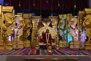 A seated man in a rust-red robe and pink-and-white flower garland sits on an ornate gold throne with a cobra hood carved above it, on a stage with golden lion-headed pillars, a red curtain and hanging brass bells. A small golden ritual vessel with a lid sits on the carpet at his feet. Colourful striped rugs cover the stage floor. The seated figure raises one hand in a gesture. The throne setting and robe colour are visible, but the individual's identity is not established from appearance, so the a person field is unsure.