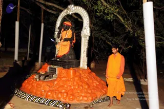 Night photo at an outdoor shrine beneath dark trees. A large black statue stands within a silver arched frame, draped in orange cloth with a red-orange head wrap and a garland of flowers around its neck. In front, an orange-painted rounded mound holds small brass oil lamps and a tray with red offerings. A young boy in orange robes stands barefoot at the right, looking down. White poles and a black-and-white dotted border surround the shrine.