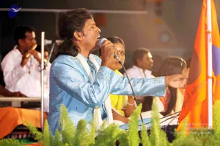 A man with long dark hair sings into a handheld microphone on a stage at night. He wears a light blue embroidered silk kurta with a white shawl draped over his shoulder, and extends his right arm outward. Behind him, a musician in white clothes plays a wind instrument, and women in yellow and orange saris sit in the background. An orange flag with white Tamil script stands at the right edge, and green fern-like foliage decorates the stage front.