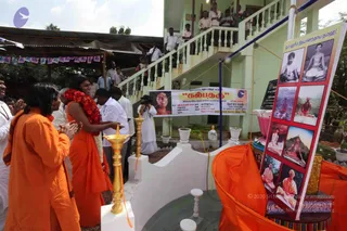 Outdoor ceremony at a pale green multi-storey building with white balustrade staircase; a crowd watches from the upper balconies. In the foreground, a woman in orange sari with a red floral garland stands at a white ritual platform with brass oil lamps and an offering of orange cloth, applauding beside a man in orange robes with raised hands. A man in white shirt and a photographer stand nearby. A tall red-framed board shows a grid of framed photographs of saints in meditation and a printed Om invocation. A banner reads Kalpataru in Tamil with event details. Bunting hangs from a canopy at left.