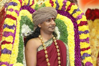 A man stands in front of a flower-decorated arch of yellow marigolds, purple and red blossoms, and green leaves. He wears a brown turban, a red dot on his forehead, a maroon garment with a gold-bordered edge, and a long gold bead necklace. Swamiji (probable): this person has long swept-back hair, a turban, a red-and-gold ceremonial outfit and a gold bead necklace, and the page indicates Swamiji attended this event. The background includes more floral decorations and a red floral display on the right.