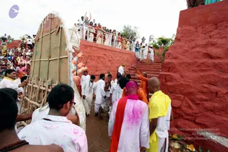 Outdoor festival procession on red-painted stone steps at a Dasara celebration. A large bamboo-framed palanquin or ratha decorated with white and gold ornaments and a clay deity murti is carried down the steps. Several men in white dhotis and white kurtas walk alongside, some wearing lanyards. A man in a yellow shawl and white clothes stands at the right, and a bald monk in an orange robe with a pink-and-orange shawl stands in the foreground, the pink dyed by colour powder. Spectators in colourful saris and white clothes line a red wall and balcony above, many raising their hands. A brass or clay offering pot sits at lower right. Speckled pink and yellow colour powder covers many of the people, and the ground is muddy.