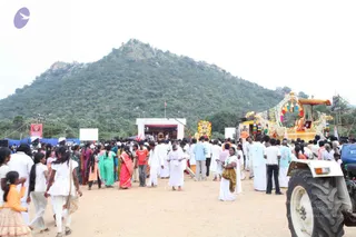 Outdoor festival ground with a dusty dirt surface and a wooded hill behind. Many devotees mill about, mostly in white dhotis and white saris, with women in green, pink and orange salwar and saris. A large white canopy stage with a banner reads Aththmanda Nithyananda... and a decorated golden palanquin or float on the right, draped with white flower garlands, orange and red tassels and a canopy, carries a seated figure. A tractor with a blue and white body is at the lower right corner. A large red banner stands at the far left. Some people in the crowd are children in white and orange dresses. No clear identification of a specific person is made from appearance.