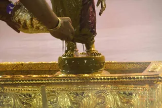 Close-up of a golden deity murti standing on a round pedestal set on an ornate gold-embossed altar, being bathed with a viscous yellow-green liquid poured by hands wearing gold bangles. The murti's bare feet and draped maroon cloth are visible, and the liquid runs down its legs onto the pedestal. Pale pink fabric forms the backdrop. Hands of a second attendant hold a gold vessel at the left edge. This is an abhishekam ritual on a statue, not a living person.