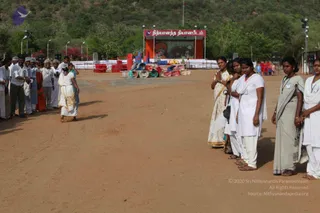 Outdoor photograph of an open dirt ground at a Nithyananda Dhyanapeetam site. A woman in a white sari with a gold border walks barefoot across the centre, away from a line of men in white shirts and dhotis on the left. On the right, a row of women in white and cream saris and white salwar stands in a line, several with hands pressed together in namaste. Behind them a stage with a red and white banner reading the Tamil name of Nithyananda Dhyanapeetam, with a large banner image, stacked red chairs, folded mats and blue and orange cloth to the side, under floodlight poles against a green wooded hillside.