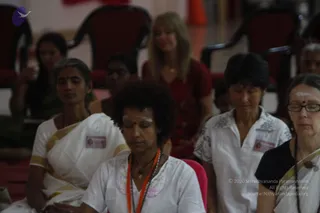 A photograph of a seated group of devotees with eyes closed in meditation at an indoor satsang hall with red chairs in the background. In the foreground, a woman with curly hair wears a white top with an orange-and-beige mala and a long pendant, with vibhuti marks on her forehead. Beside her, a woman in a white and gold-bordered sari has a red bindi. To the right, a woman in a white embroidered top and another in a black top with a white-bordered wrap and rudraksha mala sit with eyes closed. A woman in a red top with a long pendant sits in the middle background, and others sit behind them, all appearing to meditate during the talk.