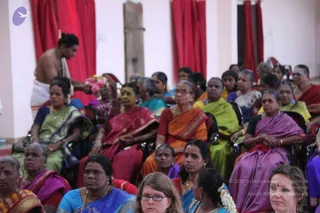 Photograph of a seated audience in a hall with red curtains and white walls, mostly women in colourful silk saris: pink, magenta, orange, green, lavender and blue. A bare-chested man in white dhoti stands at left, holding a flower arrangement with pink blooms. Several audience members wear gold necklaces, bangles and jasmine flowers in their hair. Foreground includes a woman in blue with glasses.