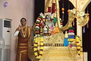 A standing man in a reddish-brown silk dress with gold-bordered yellow edges smiles and raises one hand in greeting beside an ornate golden palanquin. He wears a thick gold necklace and has a white bandage on his forearm. Behind him a deity murti is dressed in white and decorated with flower garlands in yellow, red, green and purple, with a crown and jewelled ornaments. The golden vehicle has a carved lion-head prow, bells and tassels in blue, pink and orange, and a plain white wall and an air conditioner stand on the left.