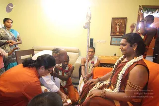 A home visit in a yellow-walled living room during a padapuja. A seated man in an orange-brown robe, white flower garlands and rudraksha malas, with a gold bangle and a gold earring, has his feet being washed by a woman in orange on the left, while a kneeling man in white holds a brass bowl. A young man in orange with a garland kneels beside them, and a young priest in white with a silver trident-topped staff stands behind. A standing young man in orange on the right holds a garland-decorated mirror, and a woman in teal and grey sari watches from the left. Framed deity murti of Venkateswara and a sofa complete the setting. Probable Swamiji: the seated figure wears long swept-back hair, rudraksha malas and a gold earring, marking him as the central figure receiving padapuja.