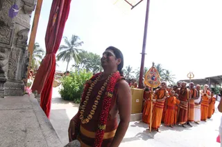 A man stands at the left edge of an open-air temple courtyard beside a carved grey stone pillar and a red curtain, smiling and gazing upward. He is bare-chested with a rust-brown dhoti-style cloth, a thick red and gold flower garland with gold beads draped over his shoulders, and a white tilak on his forehead. Behind him a line of saffron-robed sannyasis and younger boys in orange stands in a row under a palm-lined sky, with one senior man in saffron holding a yellow-and-red ceremonial standard topped with a trident.