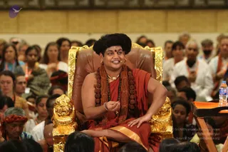 A seated man in an ornate golden throne with lion-head armrests sits at the front of a large hall, facing a seated audience. He wears a rust-orange robe with a gold-striped edge, layered rudraksha mala beads, a gold necklace, and a black swept-up hairstyle with a red bindi. He holds one hand raised in a teaching gesture while smiling. Behind him, many people sit in rows, several wearing orange and white clothes and orange delegate lanyards. A small plastic water bottle sits on a wooden lectern at right. A purple logo appears at top left. a person wearing a saffron robe with rudraksha malas and a gold necklace, seated on a throne as the discourse leader.