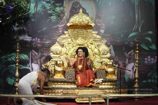 A photograph of a seated teacher on an ornate gold throne with carved lions and peacock-shaped armrests, draped in red-orange robes with a rudraksha mala and gold pendant necklace, long dark hair, and a white-and-orange forehead mark, mid-speech with raised hands and an open mouth. A standing microphone is in front. A man in white dhoti and shawl kneels at the lower left. Two figures with palms pressed together flank the throne on a painted jungle backdrop with a waterfall and a deity figure above. Ornate brass lamps stand at the sides of the stage.