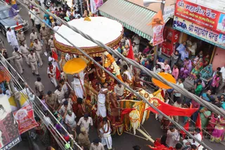 A street procession viewed from above in a crowded Indian town lane. At the centre, a decorated palanquin or ratha with a white canopy edged in gold fringe and a large orange parasol is carried by men in saffron and red garments, many bare-chested with rudraksha-style adornments, and white-clad attendants. Garlands of marigolds and white flowers drape the carriage, with a red cloth and brass kalasha-like finials. Uniformed police in khaki line the road, and red and orange flags mark the route. Devotees, many women in saris, watch from shopfronts and balconies. Shop signs read Sri Mahavir Band and display portraits. a person's identity is not established from appearance here.