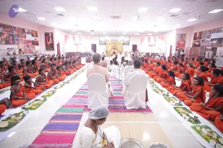 Wide view of a hall where a large group of devotees in saffron-orange robes sit cross-legged in long rows on the floor, facing a central aisle covered with colorful striped runners. Banana-leaf plates with offerings and small oil lamps lie in front of them. At the far end, a golden throne-like seat stands before a large screen. Two priests in white clothes sit on white chairs near the front, and a woman in white with a grey head covering sits in the foreground. Framed photographs and paintings line the walls. a person figure in white sits in the front row of the aisle, but his appearance is not clear enough to confirm.