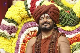 A smiling man in a rust-red turban and matching rust-orange robe sits in front of a large floral backdrop of yellow marigolds, white flowers, purple blooms and orange roses arranged in curved rows with green leaves. He wears layered rudraksha malas, a gold necklace and a red tilak on his forehead, with a small microphone clipped near his face. Swamiji (probable) appears to be the candidate here, based on the turban, saffron-toned clothing and rudraksha malas.