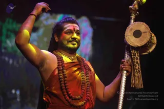 A man in an orange-red saffron garment stands on a stage under coloured lights, raising his right arm with the hand bent at the wrist. He wears a long rudraksha mala of red-brown beads and a gold necklace, with a red tilak on his forehead. In his left hand he holds a tall silver staff topped with a small damaru drum decorated with a tassel. The background is a blurred green, pink and purple projection. Swamiji (probable) appears with long swept-back hair, ornate saffron clothing, rudraksha malas and ceremonial gold necklace, matching the event page.