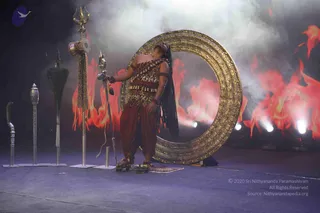 A performer stands on a blue stage dressed as a Kalabhairava-style figure, wearing a leopard-print top with a tiger-striped wrap, rust-brown dhoti-style trousers, a red sash with gold tassels, and a bead necklace. Long dark braids fall over the shoulder as the head tilts back. A tall ornate golden circular arch frames the figure, and a trident, a curved sword, a silver staff and a feathered fan stand upright on the left. Smoke and red-orange flames fill the backdrop with stage lights glowing behind.