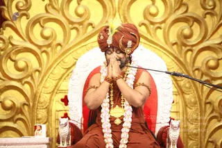A seated man in a rust-red turban with a green-and-white plume and gold ornaments sits on a white and red throne before an ornate gold scrollwork backdrop. He wears layered rudraksha malas, a white flower garland and a large gold pendant, with a microphone stand beside him. He holds his palms together in namaste with eyes closed. Two white lion figures flank the throne and a small framed photo sits at the lower left. Probable Swamiji (probable): the red turban with plume, rudraksha malas and ceremonial gold pendant are the qualifying cues. Watermarked archive photo of a satsang setting.