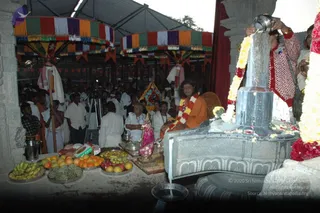 A photograph of an abhishekam at an outdoor temple pavilion under a striped canopy. A large silver-toned lingam sits on a stone yoni-peetha, with water poured over it from a metal pot held by a woman in a red patterned top. A seated figure in orange robes with a flower garland and long dark hair sits beside the lingam, and a water channel flows from the peetha. Behind them a crowd of men in white dhotis and shirts stands in the pavilion. In the foreground are trays of bananas, oranges, apples and custard apples, a brass kalasha and a small brass deity murti with a pink cloth.
