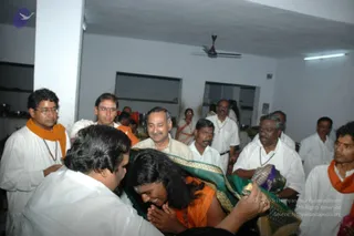 Indoor photograph in a plain white-walled room with a ceiling fan and fluorescent light. A long-haired man in an orange cloth and beaded mala, his hands pressed together, bows his head low while a white-clad man in front of him leans in and holds a green-and-gold embroidered shawl across his back. Around them, several men in white shirts and white dhotis, some wearing orange scarves and rudraksha or beaded necklaces, look on; a man in a cream shirt smiles at the center. Steel vessels and a dark cabinet stand along the left wall.