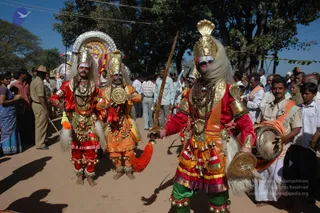 Outdoor photograph of a street procession in Karnataka with three performers in elaborate costume on a dirt road under large trees. Foreground performer holds a long sword and wears a gold crown, red and gold bodice, rudraksha-style beaded necklaces, bells at the ankles and a round gold shield at the hip. Two companions beside him wear gold crowns, white fringed headdresses, orange and red tunics and bells, one holding a round shield. A drummer in white at right holds a large drum. Behind them, a crowd of onlookers in white shirts, saris and a uniformed police officer, with a decorated float or chariot and bunting in the background.