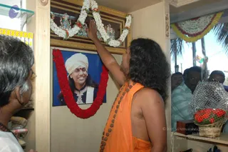 A photograph inside a shop interior showing a bare-chested man with long dark hair, wearing an orange saffron cloth and a long rudraksha mala, reaching up to hang a white and red flower garland on a framed image. Above the frame sits a small gilded deity murti set among white flower garlands. The framed portrait below shows a smiling man in a white turban with a red tilak, a dark jacket and ornate embroidered collar, draped with a red flower garland. To the left, an older man in a white shirt watches. Behind them, several onlookers stand in a doorway, and a basket of red flowers sits on a shelf at right beside a palm-decorated entrance with bunting. The scene suggests a shop opening or blessing ceremony.