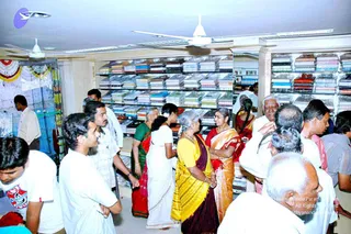 A photograph of a crowded textile shop interior with shelves stacked with folded shirts and fabric bolts in many colours. Ceiling fans turn overhead. Several men in white shirts and a few women in sarees stand near the centre of the room, some in conversation, one woman in a yellow saree and maroon border and another in a red and gold saree. A garlanded display area with marigold garlands is visible at the left edge.