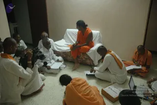 Indoor photo of a seated gathering on a plain tiled floor against a pale wall. A woman in an orange sannyasi-style garment with dark hair pinned up sits cross-legged on a white-draped couch, facing a group. Around her, men in white dhotis and shirts and men in orange robes sit on the floor; one man in white with a shaved head writes on a notepad, another in orange writes in a notebook, and a man in white holds a microphone. A videographer in white with a camcorder films at left, and a laptop sits on the floor at lower right. A small box with a logo lies in the foreground.