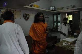 A photograph taken inside a small plain room with white-washed walls, a wooden bench and a counter covered with a patterned cloth holding an open ledger and papers. A man with long swept-back dark hair wears a flowing saffron-orange robe and speaks while gesturing with his right hand; the appearance is consistent with the Swamiji (probable) candidate on the page, a long-haired figure in orange robes, and this is an in_person candidate. A man in a white shirt and white dhoti stands at the counter writing, another man in a light striped shirt stands at the right edge, and a partly visible person in white stands at the left. Wall items include a calendar, a heart-shaped framed decoration, a small framed picture and a few brass figurines on a shelf near the doorway.