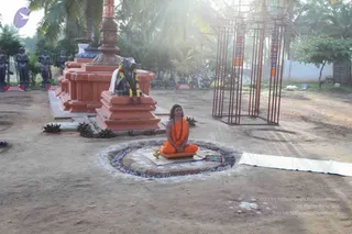 Outdoor photo in a sandy courtyard beside a palm-fringed grove. A single seated person in an orange-saffron robe with a rudraksha mala around the neck sits cross-legged on a mat within a circular stone-lined platform decorated with white rangoli. Behind is a tall orange and red pillar and a red-brick base holding a dark bull statue garlanded with flowers. Several dark stone figures stand on a platform at left, and a tall red metal frame structure stands at right with orange banners. Sunlight flares through the trees. Small placards sit at the bases of the statues. The seated person's appearance is consistent with Swamiji (probable), given the saffron robe, long hair and rudraksha mala, though identity is not certain.