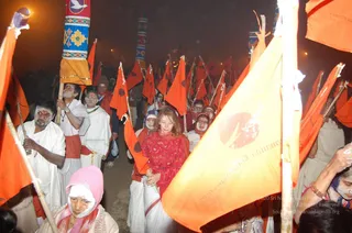 Night photograph of a procession at the Kumbh Mela with many devotees carrying tall bamboo poles topped with saffron-orange flags. Several men in plain white dhotis and white shawls walk with faces smeared with white ash, and some wear white headcloths. A woman in a red blouse and white sari walks in the centre holding hands with another devotee, surrounded by orange flags bearing a red emblem and Devanagari lettering. Blurred lamps and dark sky form the background. The people count is more than twenty but this is an estimate from the visible crowd.