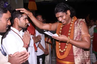 A photo of a satsang-style blessing. A man with long swept-back hair, a red-brown wrap and orange-beaded necklaces with large gold bead ornaments, plus gold armbands, places his right hand on the head of a young man in white kurta with a rudraksha mala and red thread. The blessing figure holds a folded white paper in his left hand and smiles. The young man stands with palms pressed together in namaste. Other people in white and red garments, a man in a grey shirt and a woman in a green sari stand behind, with a crowd in the dim hall. The blessing figure matches Swamiji (probable) by long hair, orange beads, gold ornaments and wrap, with the blessing role.