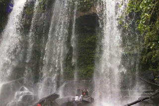 A photograph of a powerful multi-tiered waterfall cascading down a moss-covered rock face surrounded by lush green forest. Several people stand on dark wet boulders at the base of the falls, partly in the spray. One figure in white clothing and another in orange and dark clothing stand close together on a rock, while others in dark clothes stand nearby. Water mist fills the scene and tree branches frame the right edge.